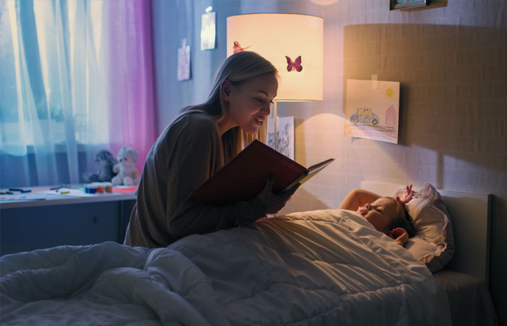 Frau liest Kind am Bett eine Gute-Nacht-Geschichte vor – Symbol für Geborgenheit und empathische Grundpflege bei Kindern
