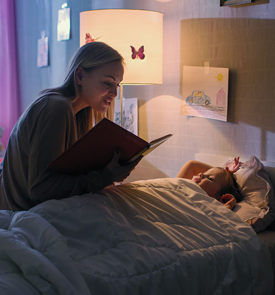 Frau liest Kind am Bett eine Gute-Nacht-Geschichte vor – Symbol für Geborgenheit und empathische Grundpflege bei Kindern